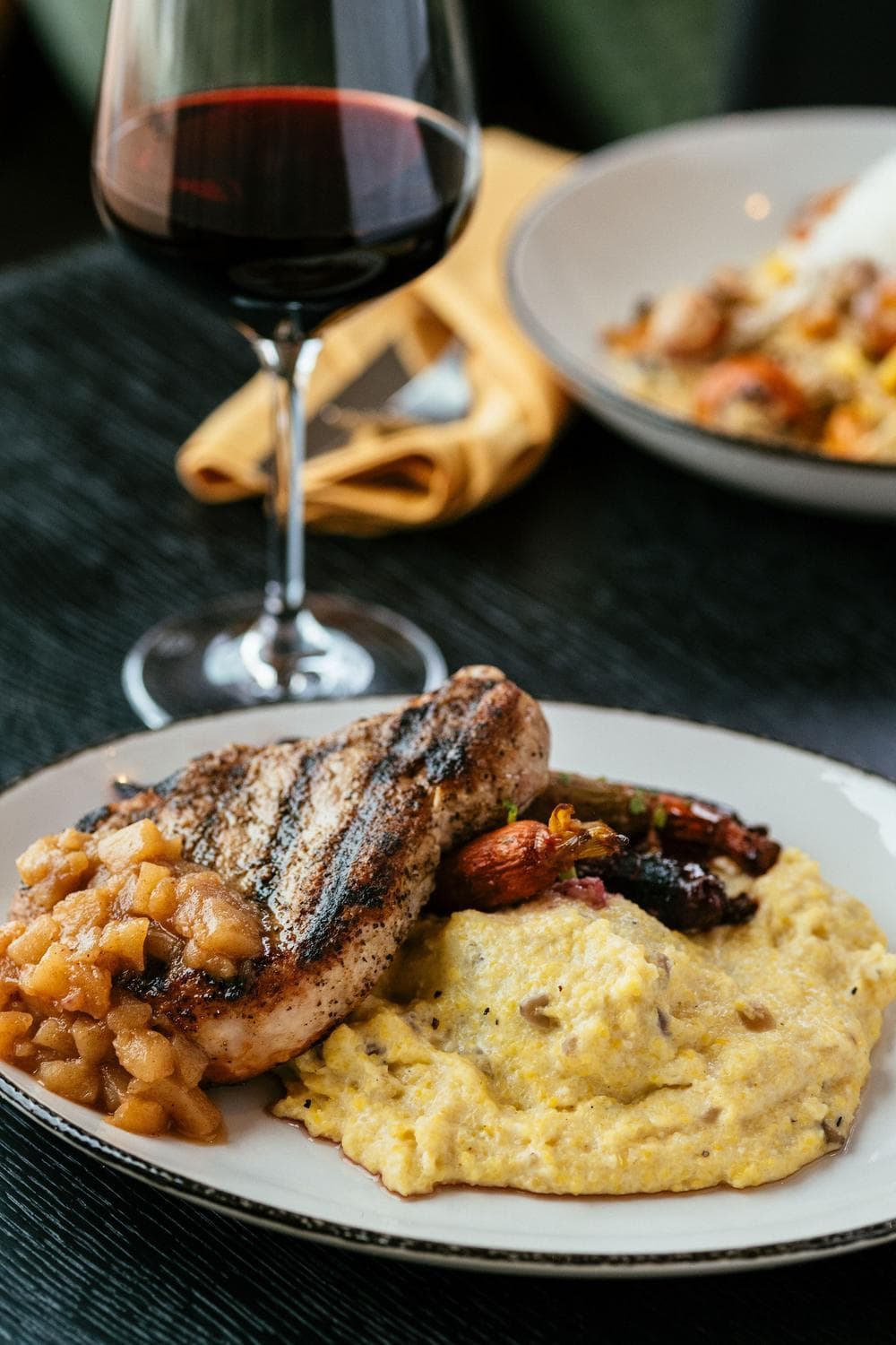 A plate with grilled pork chop topped with apple chutney, creamy polenta, and vegetables, next to a glass of red wine on a dark table. Another similar plate is blurred in the background.