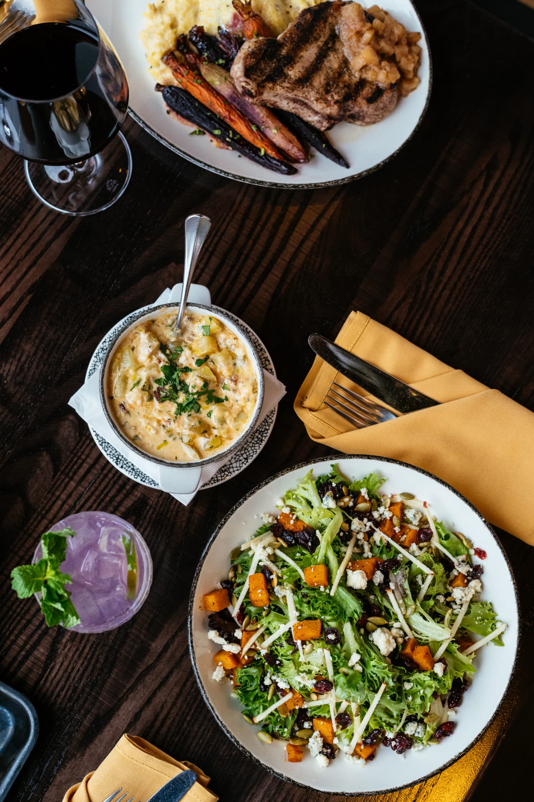 A table with a grilled meat dish, creamy side, fresh green salad with squash and feta, a glass of red wine, and a purple cocktail garnished with mint, all atop a dark wooden surface.