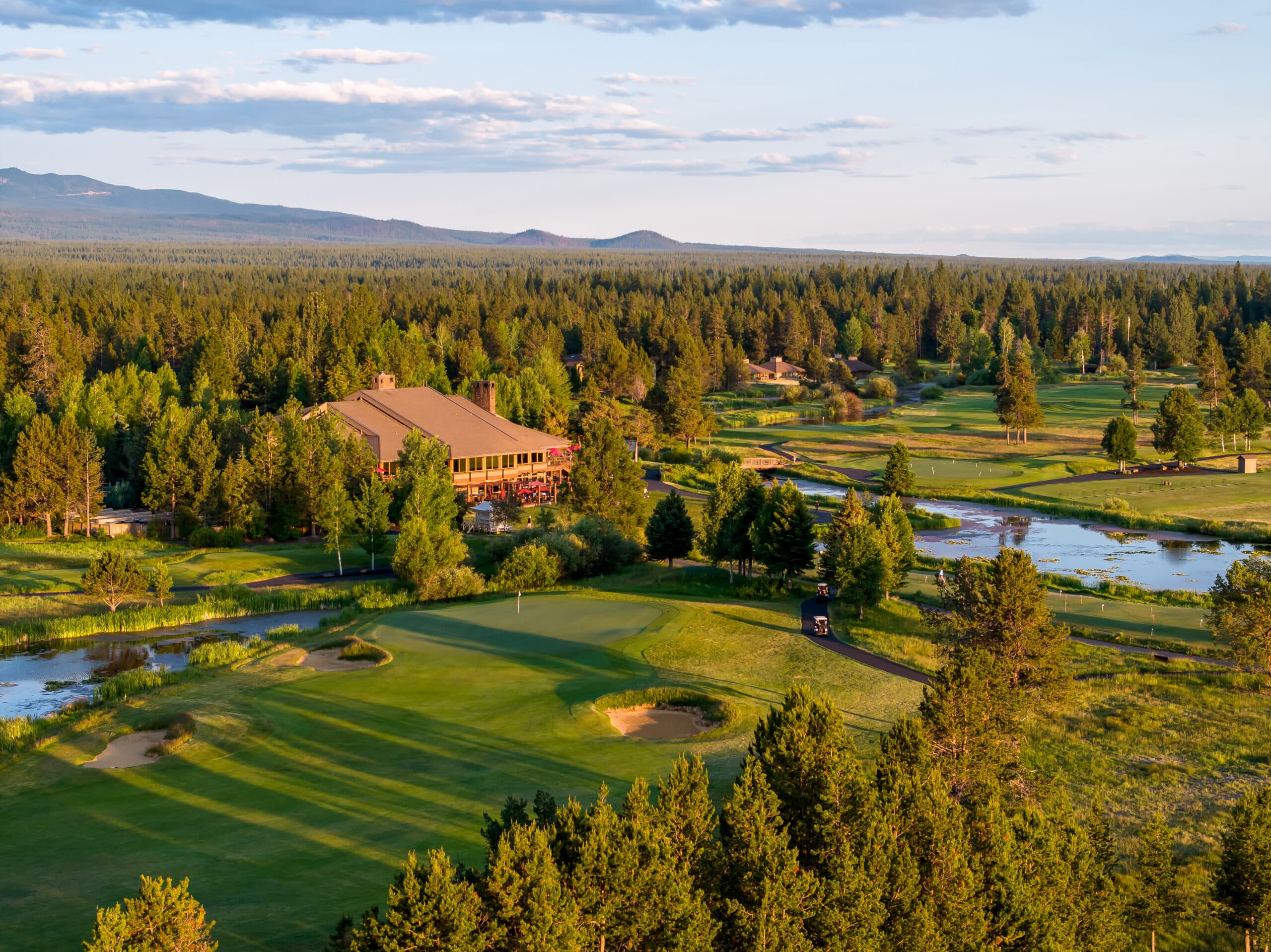 Aerial view of a golf course with a clubhouse surrounded by trees, green fairways, sand traps, small ponds, and distant mountains under a partly cloudy sky.