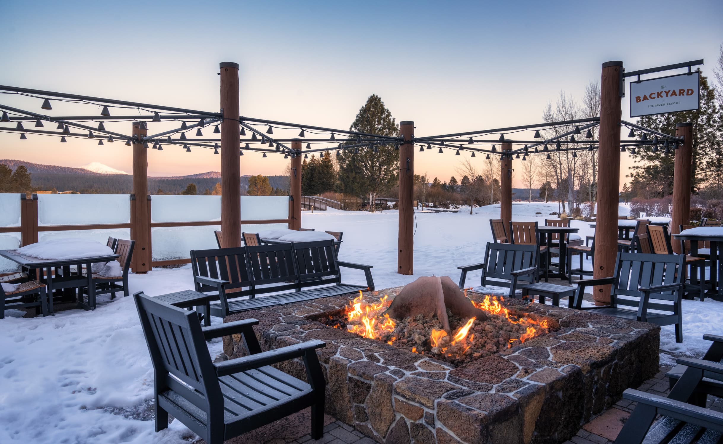 Outdoor patio with wooden chairs around a stone fire pit, surrounded by snow and string lights, overlooking trees and mountains under a clear evening sky. A sign reads Backyard on a wooden post.