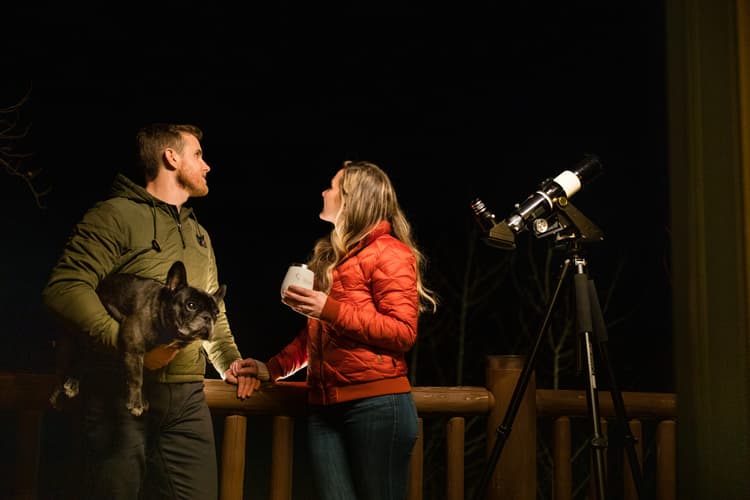 A man holding a dog and a woman with a mug stand on a wooden deck at night next to a telescope, looking up at the sky together.