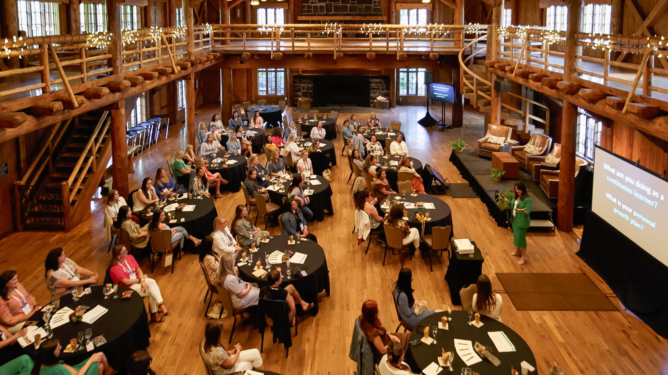 Large group of people sit at round tables in a rustic wooden hall, listening to a speaker at the front near a large screen. String lights and natural light from windows create a warm atmosphere.