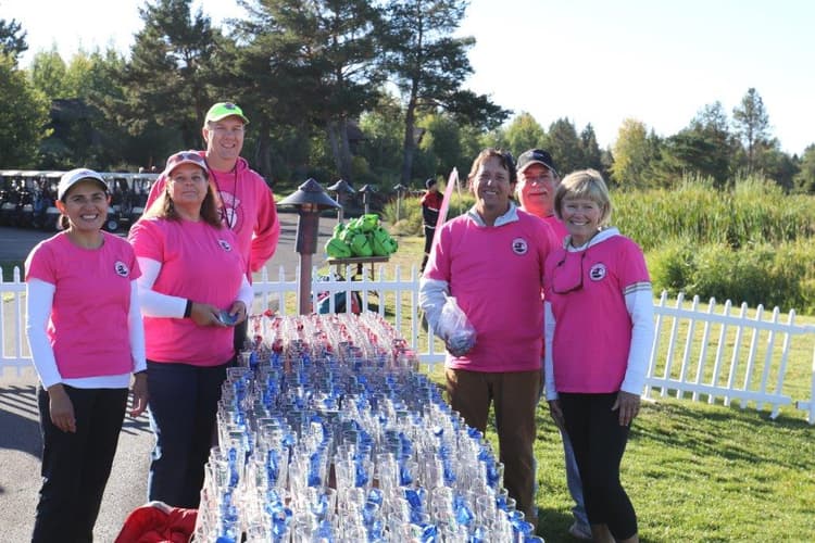 Six people in pink shirts stand outside behind a table filled with water bottles, smiling at the camera. White picket fencing, greenery, and golf carts are visible in the sunny background.