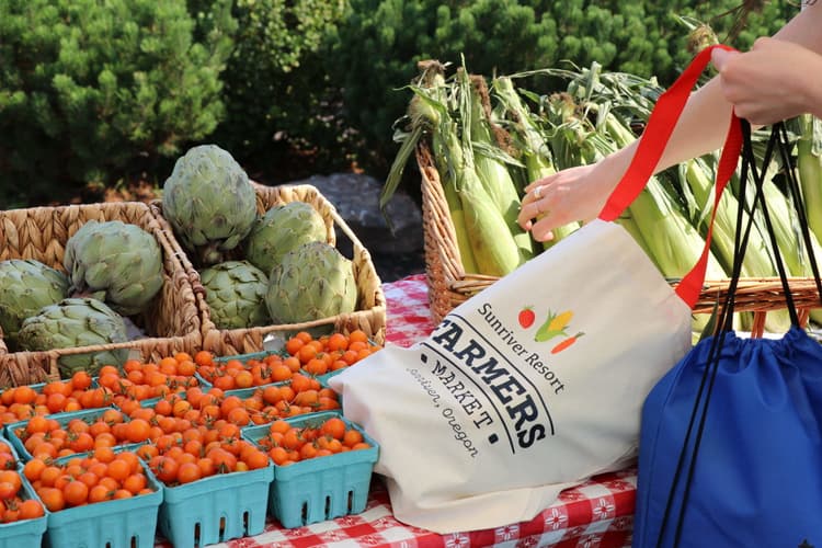 A person places corn into a reusable Sunrise Resort Farmers Market tote bag at a stand with artichokes, cherry tomatoes, and corn, on a red checkered tablecloth.