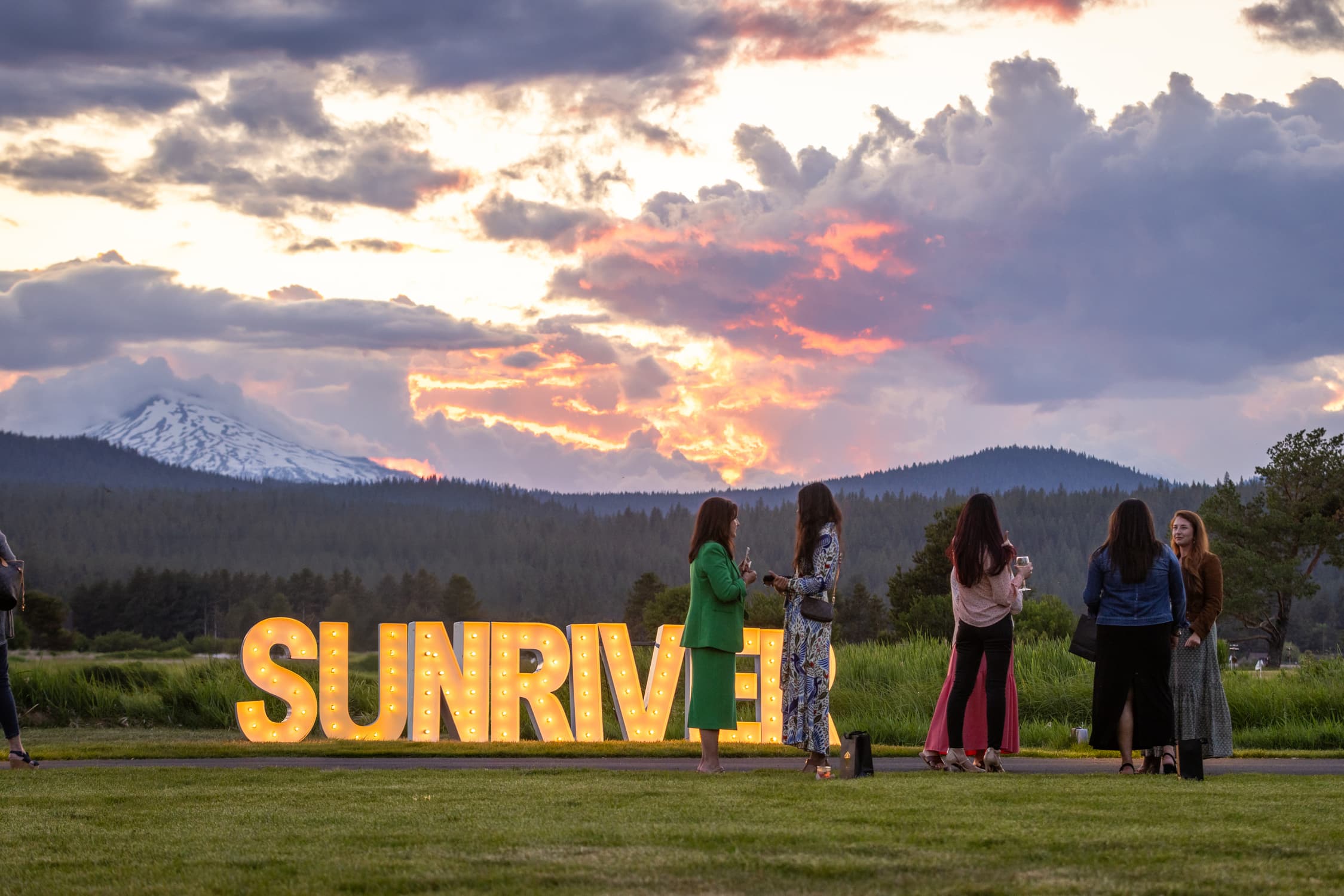 People stand and talk on grass at sunset in front of large, illuminated letters spelling SUNRIVER, with mountains, trees, and a cloudy sky in the background.