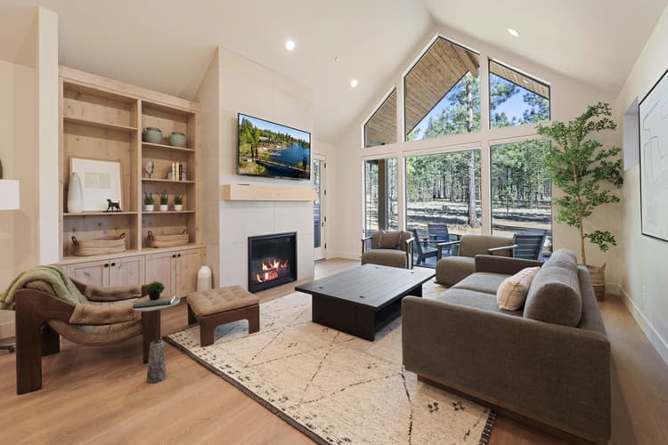Modern living room with large windows, gray sofas, a fireplace, wall-mounted TV, built-in shelves, and a scenic forest view outside. Light wood floors and a neutral rug complete the cozy, natural look—where time as an ingredient is thoughtfully woven into every detail for an inviting and timeless atmosphere.