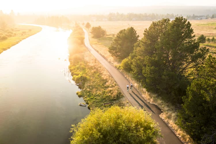 A paved path runs along a river at sunset, bordered by grass and trees. Two cyclists ride along the path, casting long shadows in the golden light.