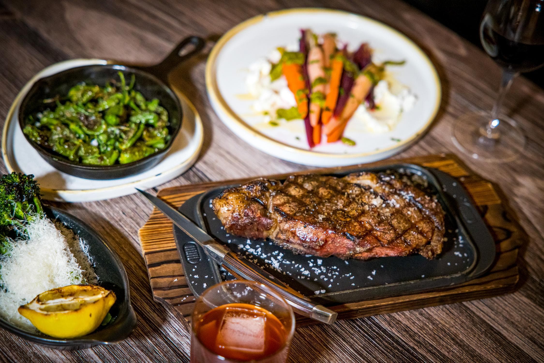 A grilled steak on a platter with a knife, served with broccolini and lemon, sautéed green peppers, a carrot and cheese salad, a glass of red wine, and a cocktail on a wooden table.