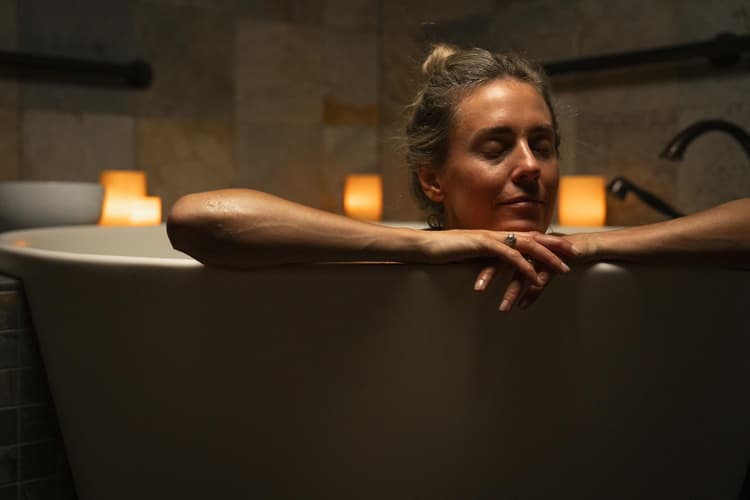 A woman relaxes in a bathtub with her eyes closed, arms resting on the edge. Soft candlelight glows behind her, creating a calm and peaceful atmosphere.