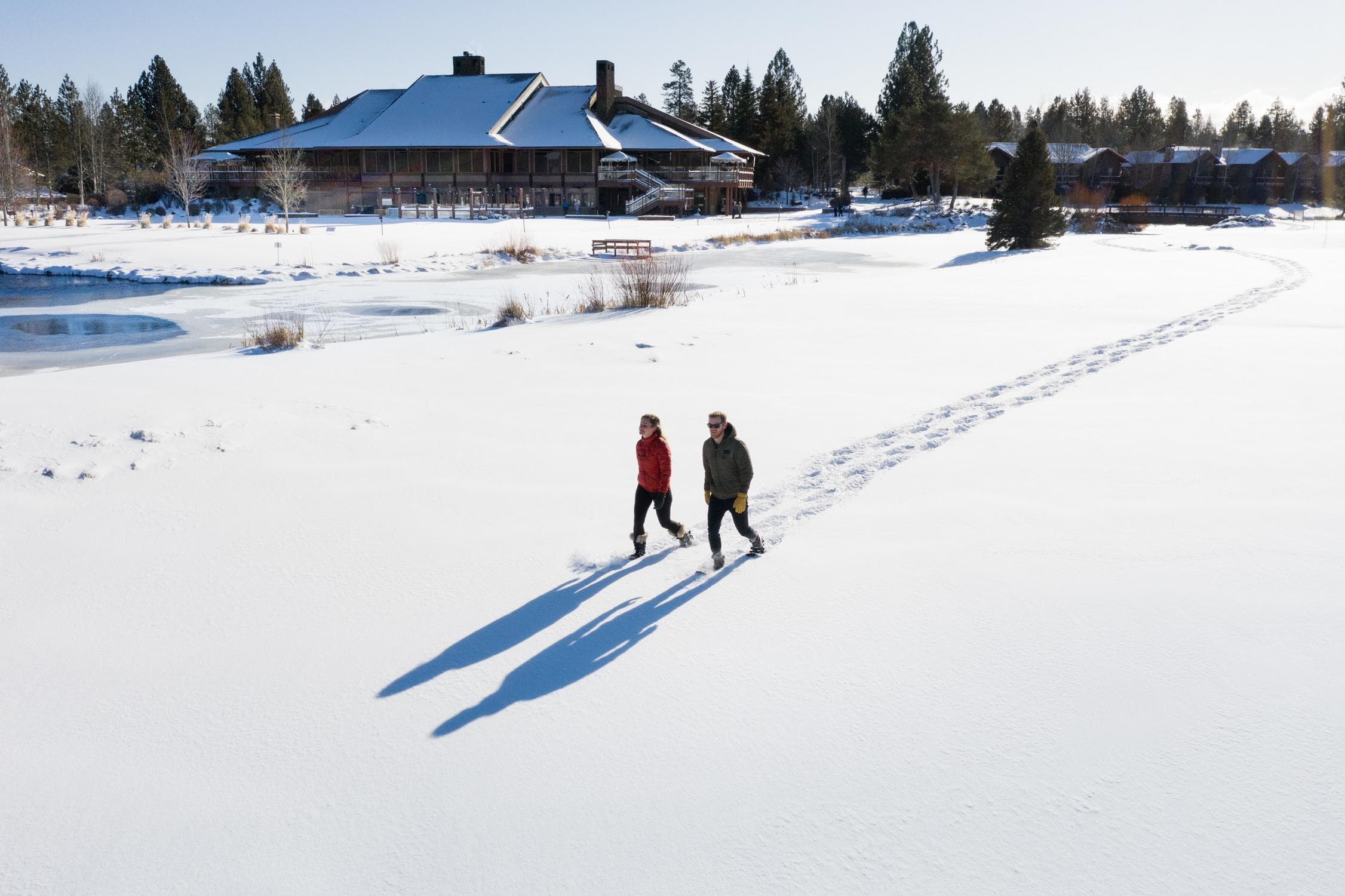 Two people walk in fresh snow, leaving footprints behind. Shadows stretch long on the white field. A large house, trees, and a small frozen pond are in the background under a clear blue sky.