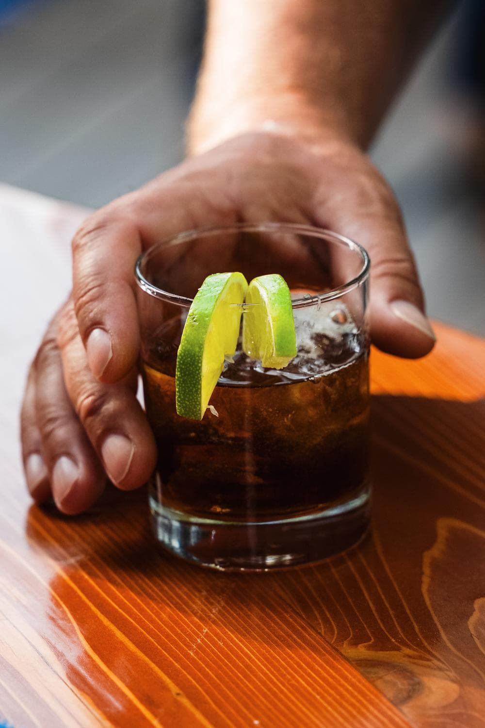 A hand holding a glass of dark soda with ice, garnished with two lime wedges, on a wooden table.