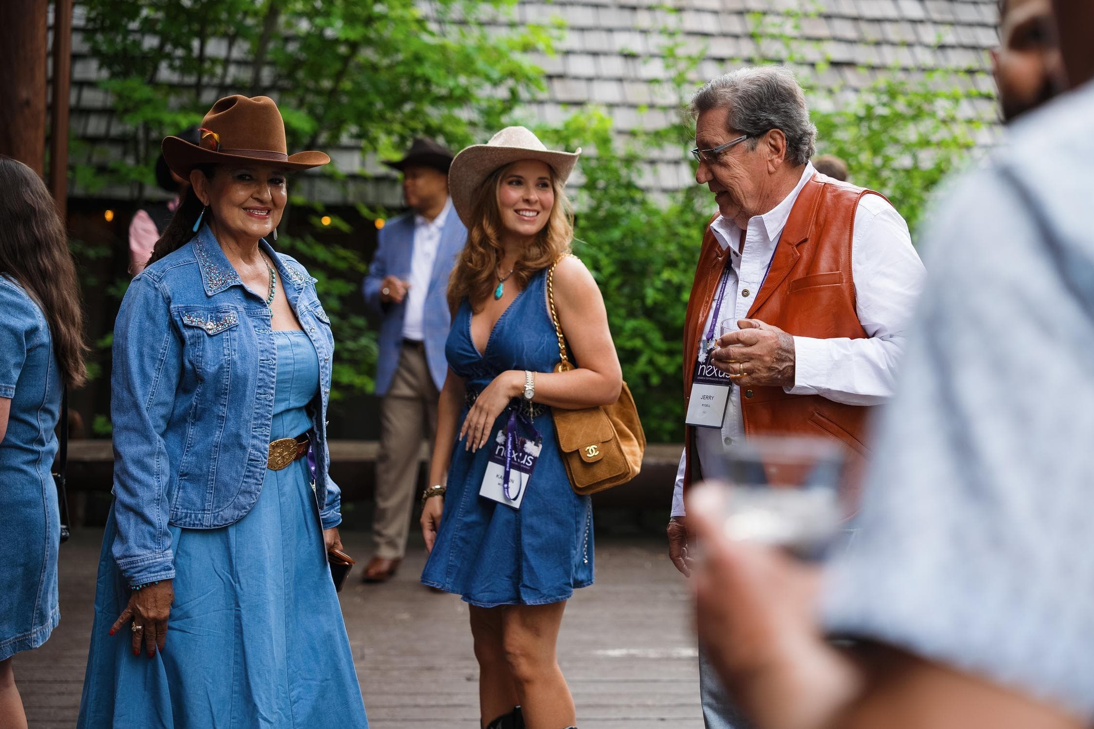 Three people in western attire—two women in denim dresses and cowboy hats, and a man in a leather vest—smile and converse outdoors at a casual event. Trees and a wooden building are in the background.