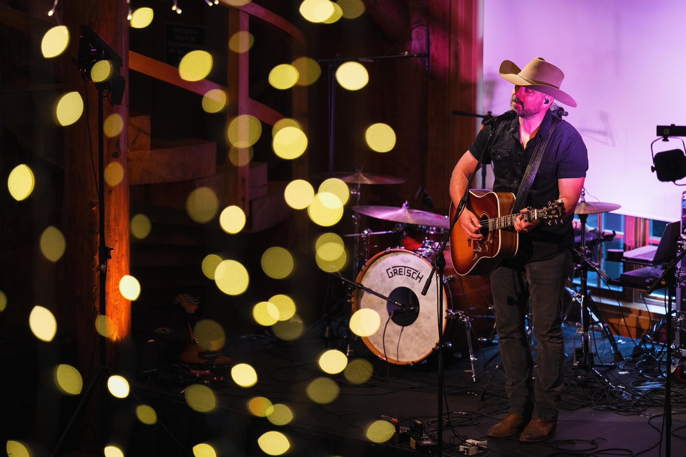 A musician in a cowboy hat plays guitar and sings on stage, with a drum set behind him. Blurred yellow string lights hang in the foreground, creating a warm, festive atmosphere.