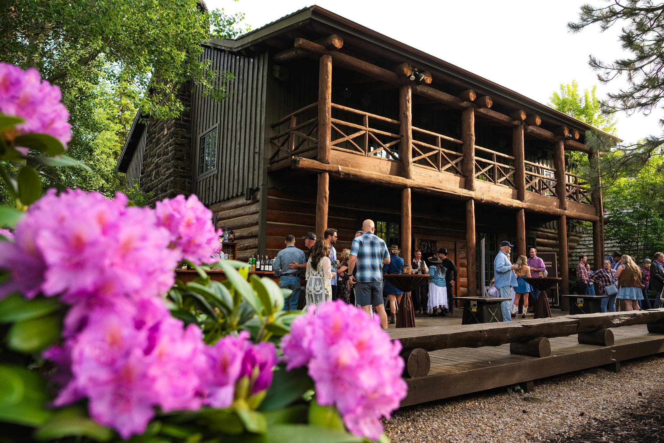 People gather and socialize on the wooden porch of a rustic log cabin, surrounded by lush greenery and blooming pink flowers in the foreground.